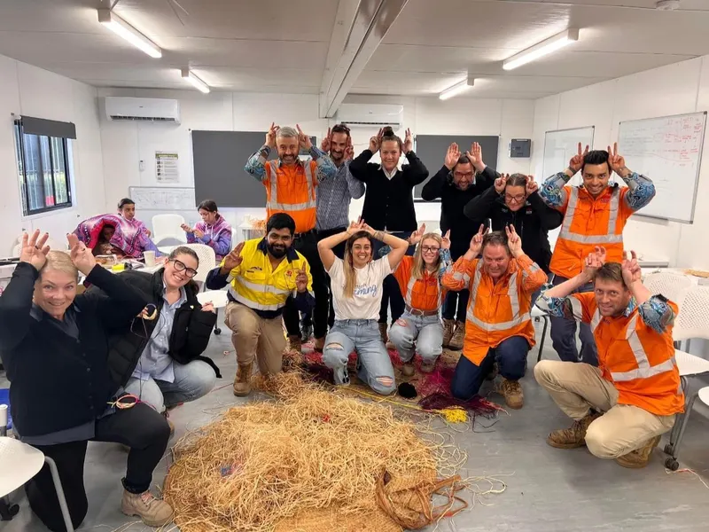 A full workshop group in a training room, about half in hi-vis, gathered around a pile of raffia and woven fibre — everyone making the emu-ears sign. Renae Lamb is front row centre